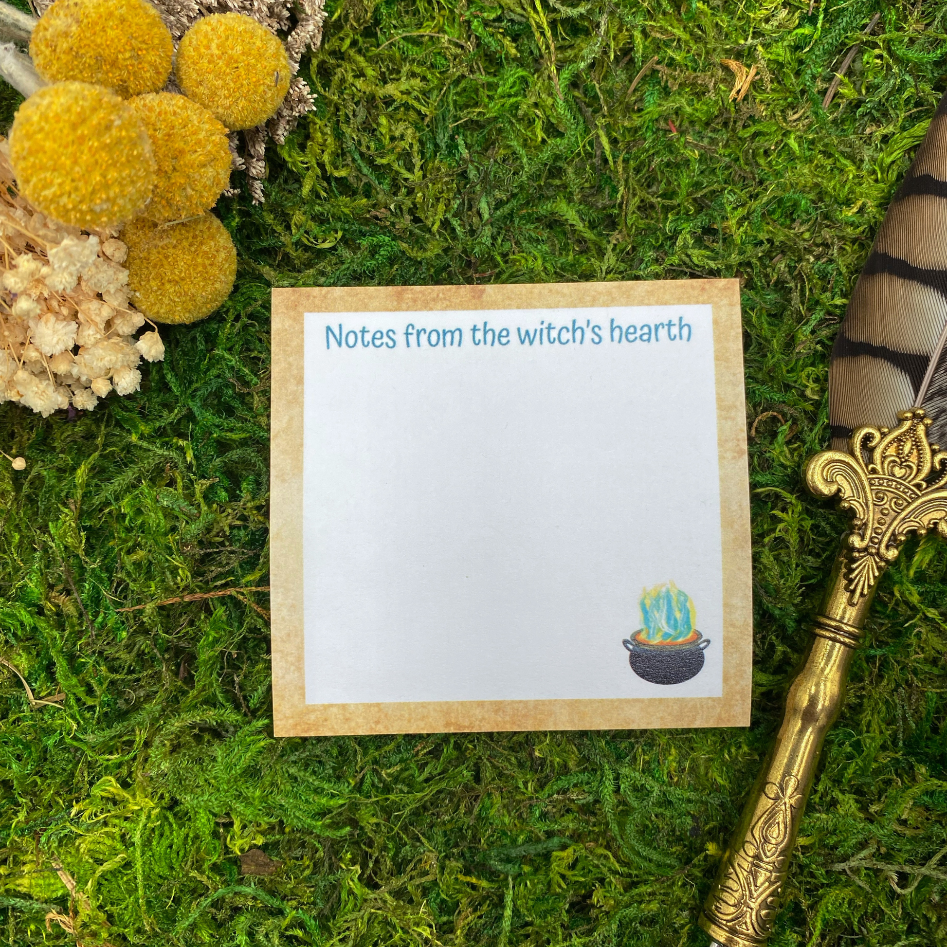 Square sticky note pad with a parchment-textured border, a small illustrated cauldron with green and yellow steam, and the words "Notes from the witch's hearth," resting on moss next to dried yellow and white flowers and a gold quill pen.