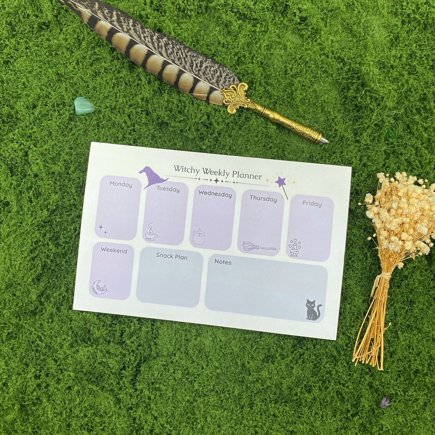 Witchy Weekly Planner notepad on moss mat with gold feather quill pen, green heart crystal, and cream dried flowers