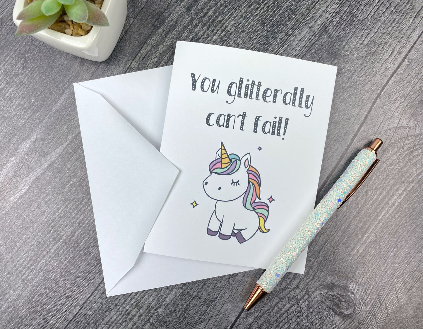 Greeting card that reads "You glitterally can't fail!" with an illustration of a small pastel unicorn, propped against its envelope on a grey wood surface next to a glitter pen and a succulent.