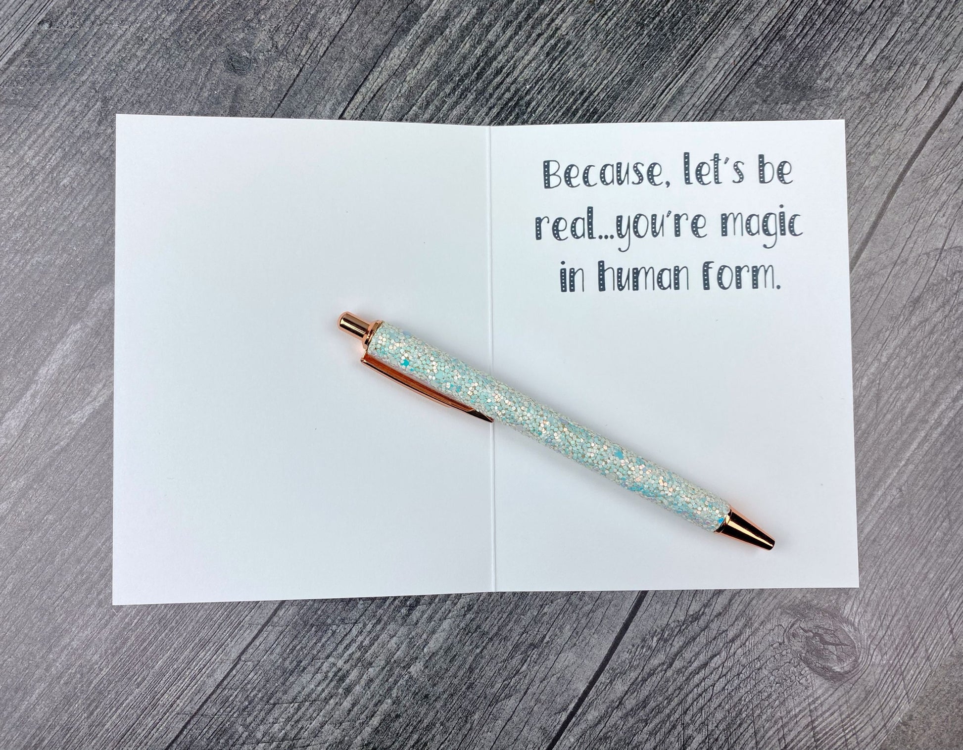 Open greeting card showing the printed message "Because, let's be real...you're magic in human form," lying on a grey wood surface with a glitter pen resting across the card.