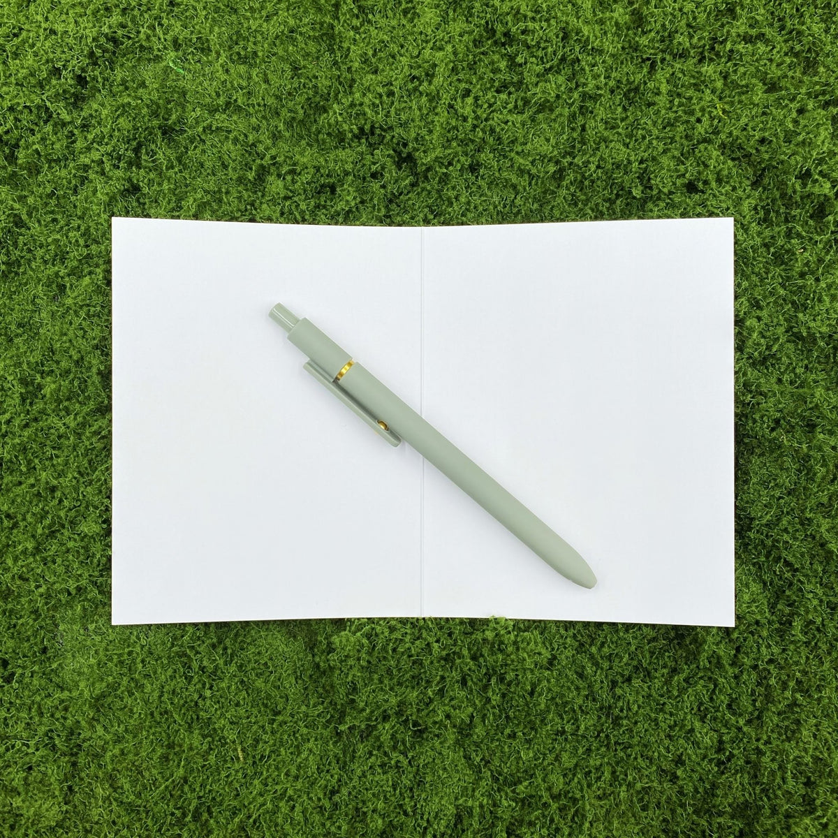 Card opened to show the blank white interior, ready for a handwritten message, with a sage green pen laid across the crease on a mossy background.
