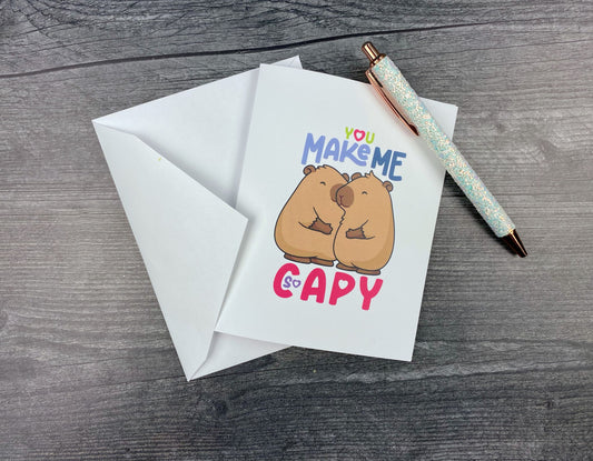 Greeting card that reads "You make me so capy" with two cuddling capybaras, propped against its white envelope on a grey wood surface next to a white glitter pen.