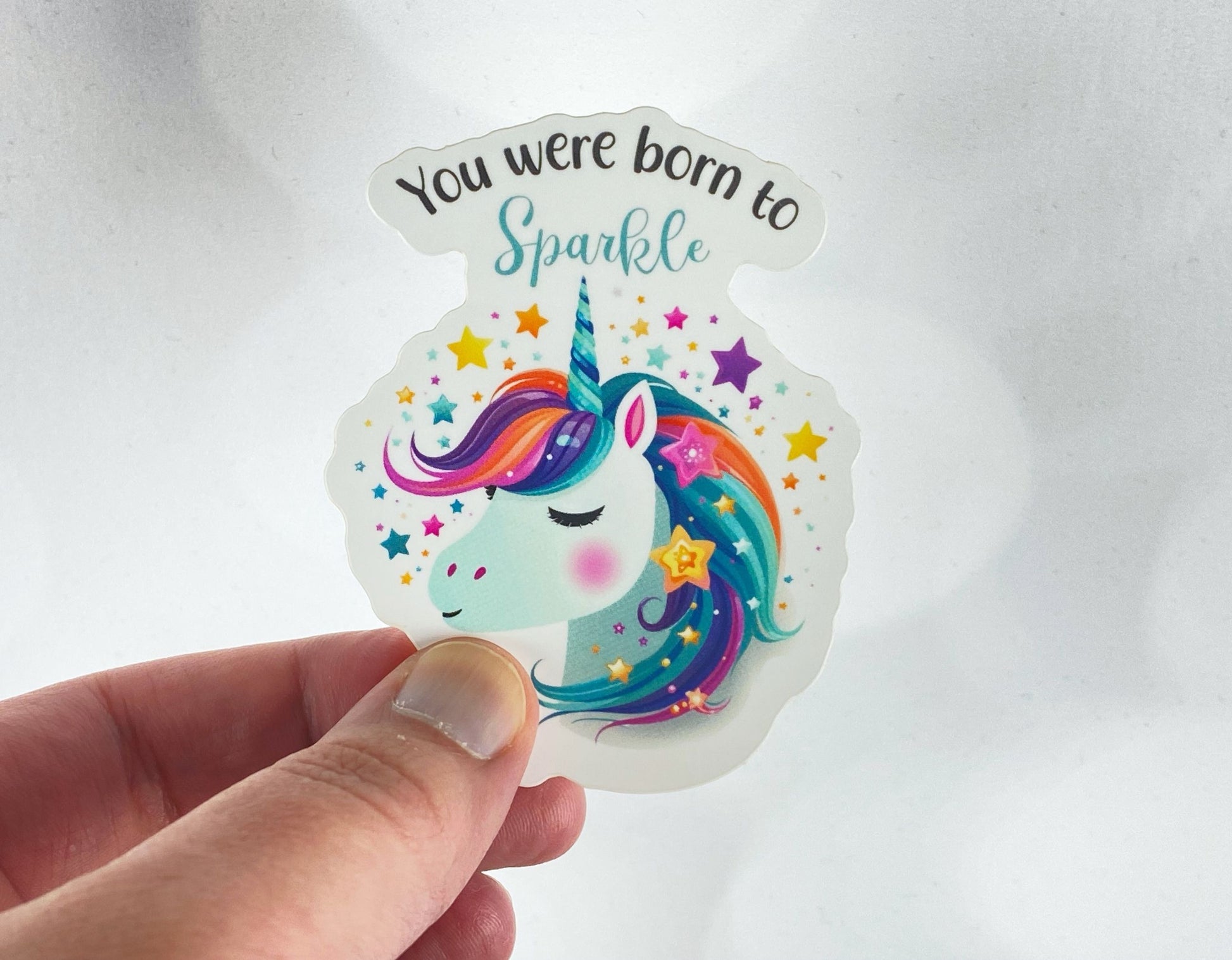 A hand holds a sticker illustration of a rainbow-maned unicorn surrounded by colorful stars, reading "You were born to Sparkle," against a white background.