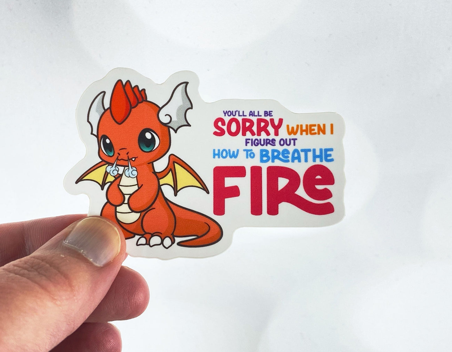 Die-cut vinyl sticker held between fingers, showing an orange baby dragon puffing smoke, with the text "You'll all be sorry when I figure out how to breathe fire," on a white background