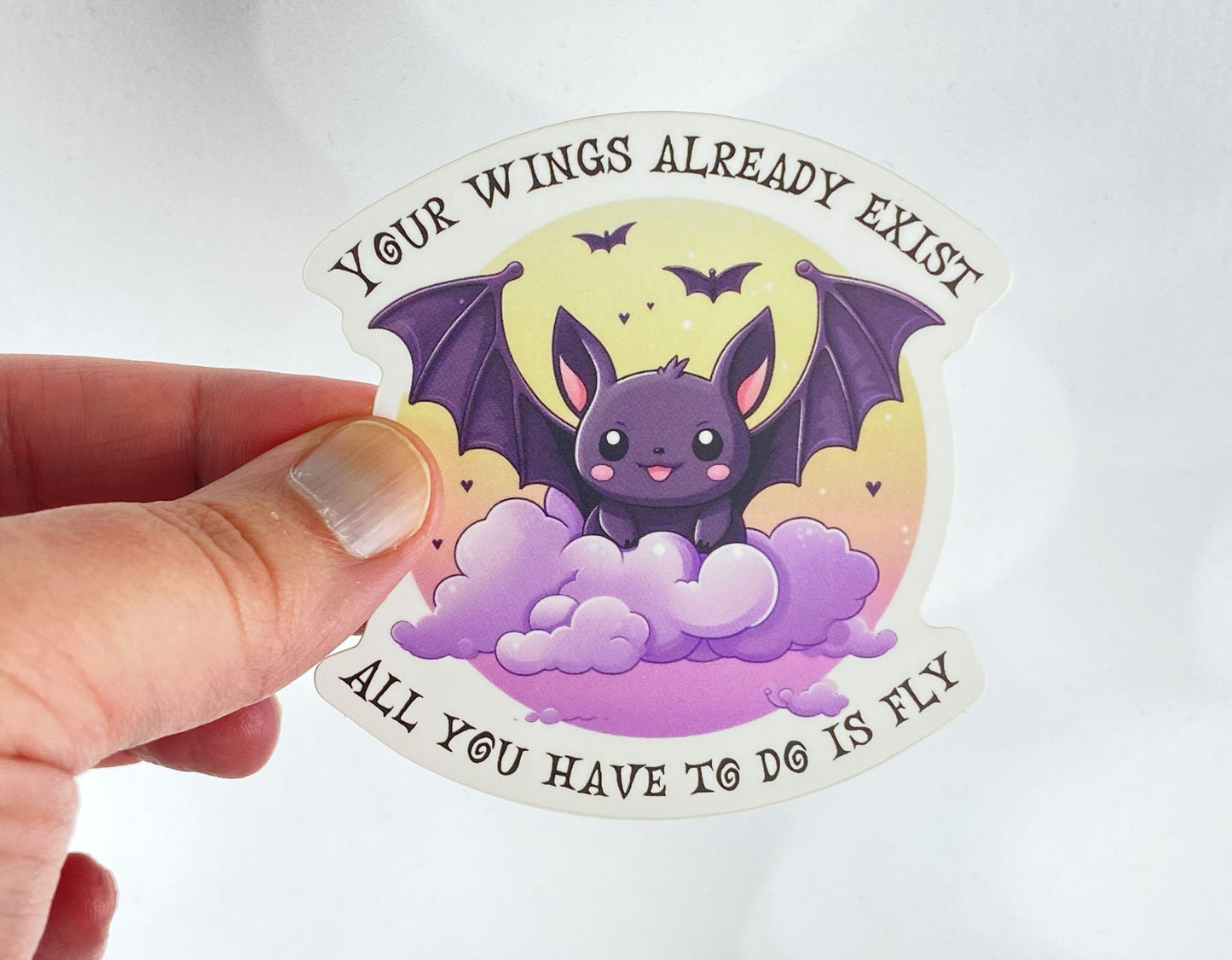 A hand holds a sticker illustration of a smiling purple bat perched on clouds under a moon, reading "Your wings already exist, all you have to do is fly," against a white background.
