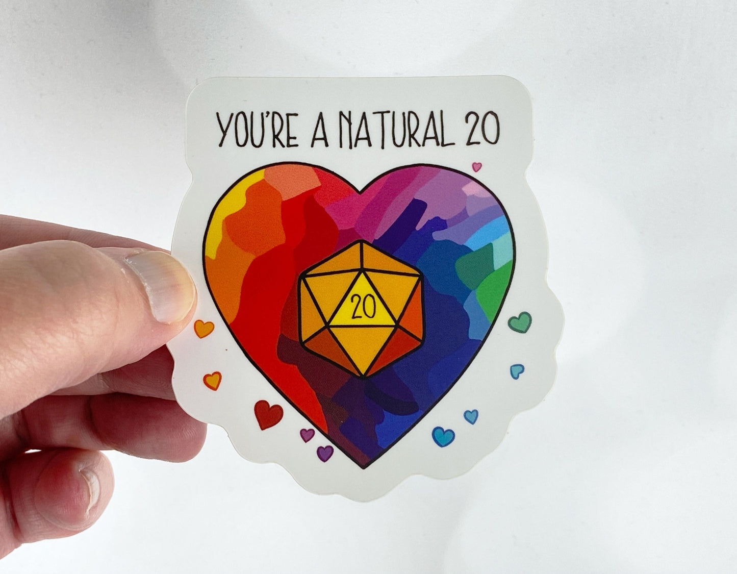 Die-cut vinyl sticker held between fingers, showing a rainbow heart with a glowing d20 in the center, with the text "You're a natural 20," on a white background