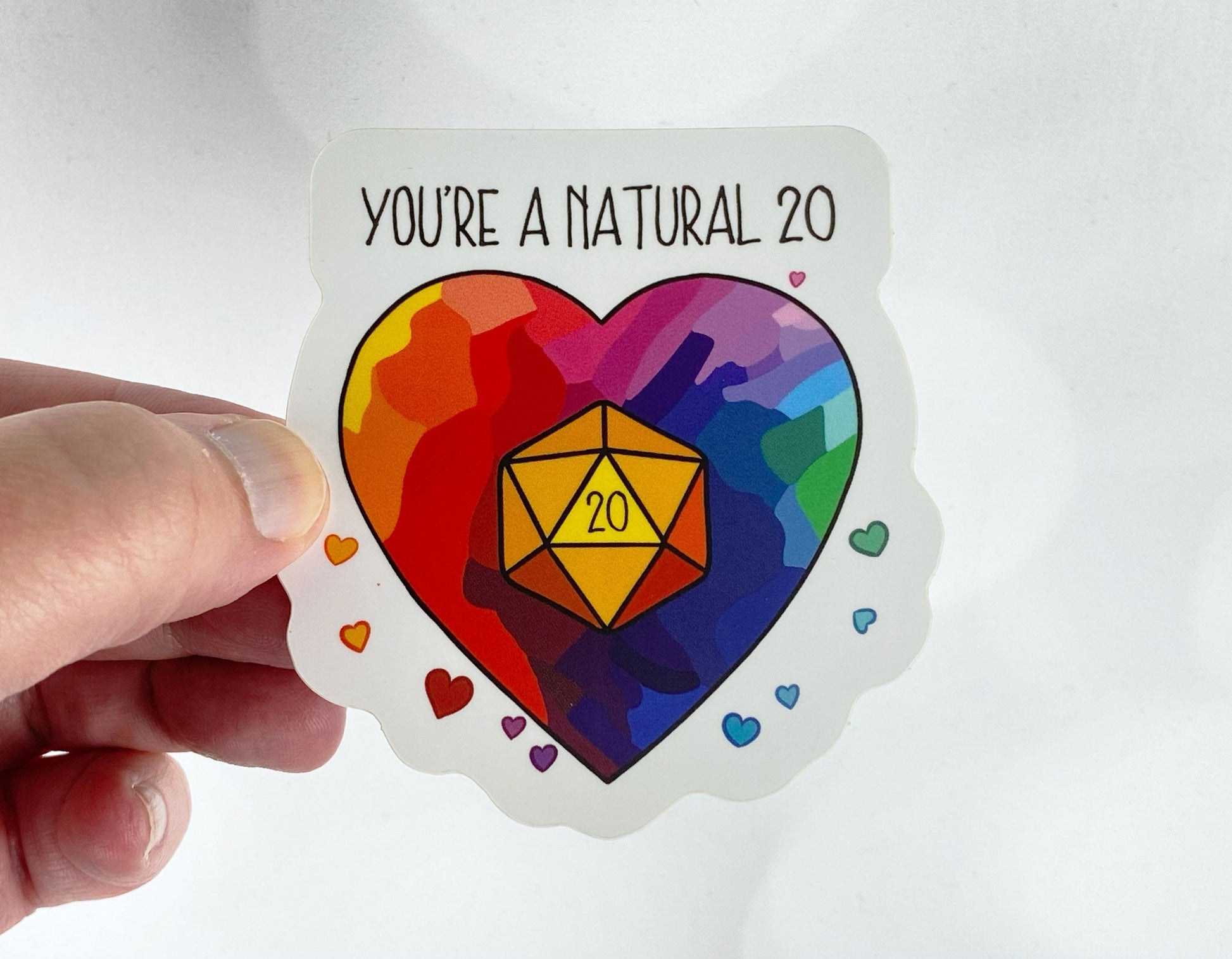 Die-cut vinyl sticker held between fingers, showing a rainbow heart with a glowing d20 in the center, with the text "You're a natural 20," on a white background