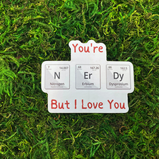 A sticker reading "You're...but I love you" with three periodic table elements (Nitrogen, Erbium, Dysprosium) spelling out "N-Er-Dy," resting on artificial moss.