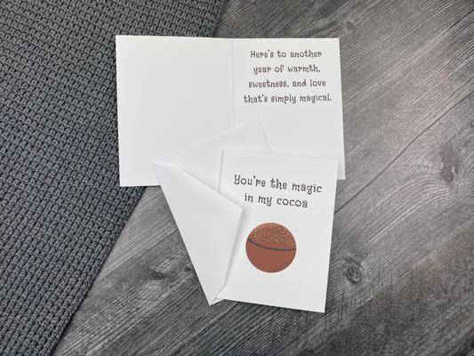 Greeting card lying open on a grey knit blanket and wood surface, showing the front with "You're the magic in my cocoa" and an illustration of a drizzled hot cocoa bomb, next to the inside message "Here's to another year of warmth, sweetness, and love that's simply magical," with the envelope tucked between the front and inside pages.