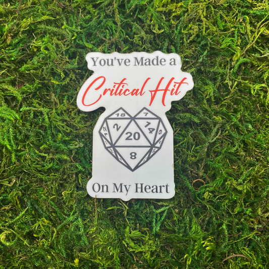 Die-cut matte sticker featuring a d20 die shaped like a heart with the number 20 in the center, with the text "You've Made a Critical Hit" in red script and "On My Heart" in gray below, displayed on a mossy green surface.