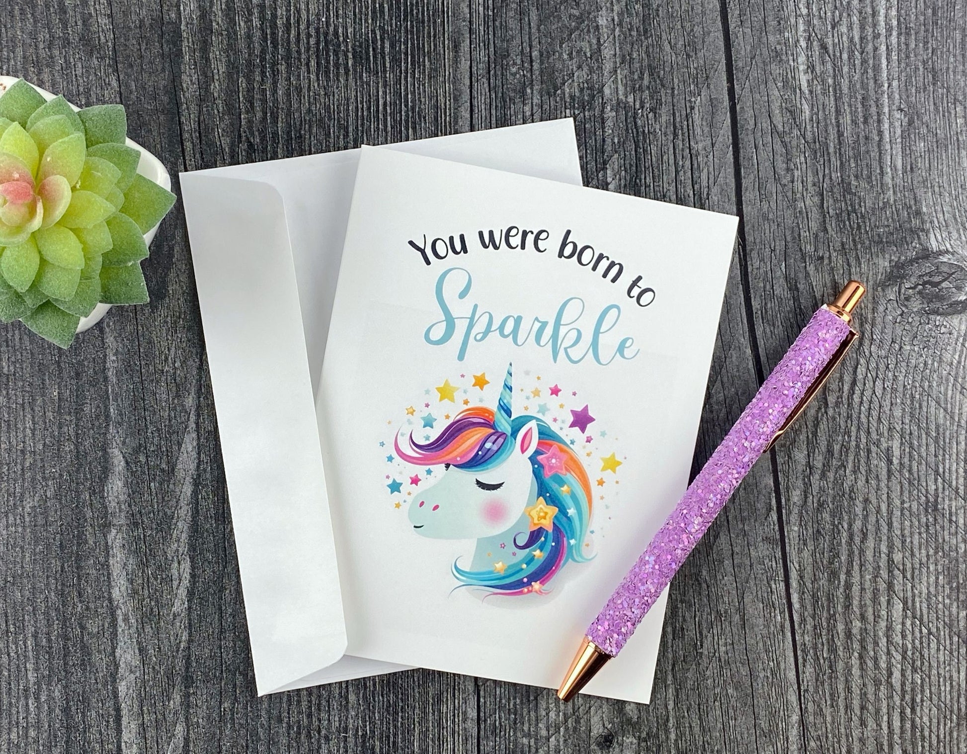 Greeting card and white envelope, featuring an illustrated rainbow-maned unicorn surrounded by stars, reading "You were born to sparkle," resting on a grey wood surface next to a purple glitter pen and small succulent.