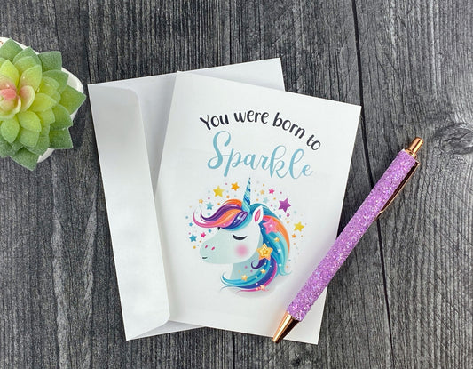 Greeting card and white envelope, featuring an illustrated rainbow-maned unicorn surrounded by stars, reading "You were born to sparkle," resting on a grey wood surface next to a purple glitter pen and small succulent.