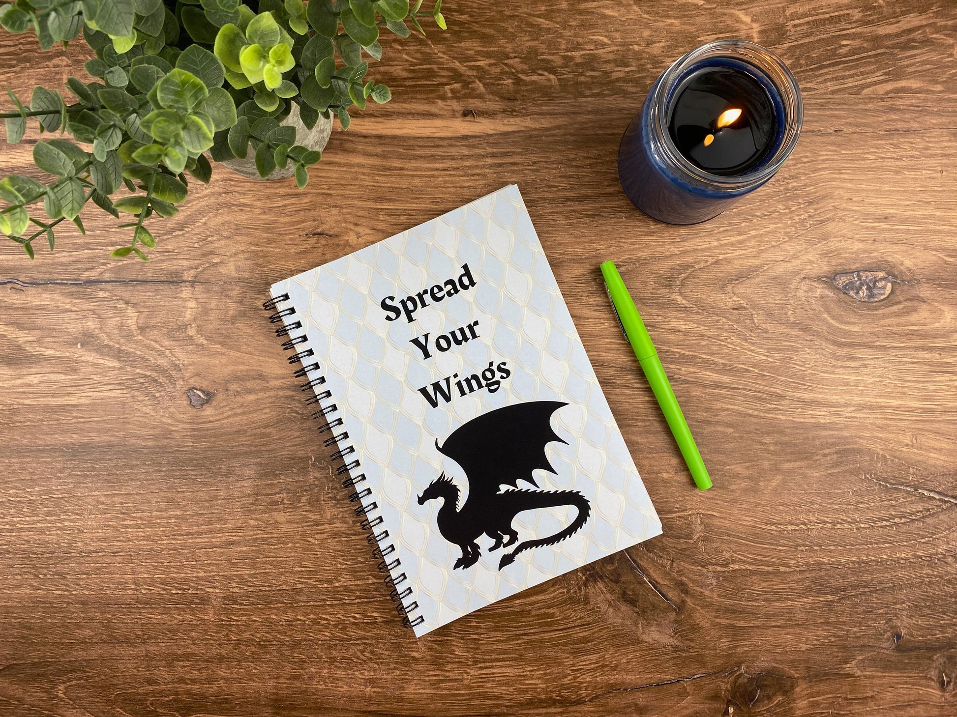 Spiral notebook cover with a pale blue dragon-scale pattern, a black silhouette dragon, and the words "Spread Your Wings," resting on a wood table next to a lit candle, a green pen, and a small potted plant.